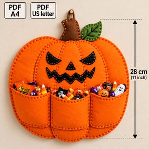 May include: An orange felt pumpkin wall hanging with a brown stem and green leaf, featuring a black stitched jack-o'-lantern face and three front pockets filled with assorted candies. It measures 28 cm or 11 inches in height.