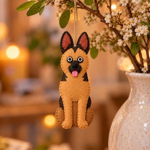 May include: A felt German Shepherd dog ornament featuring tan and black coloring, pointed ears with reddish-brown centers, white and black googly eyes, a black nose, and a small pink tongue, suspended by a thin twine loop for hanging.