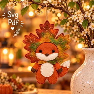 May include: A felt fox ornament featuring a large maple leaf headpiece in shades of orange, yellow, and green. The fox has a white belly, dark brown paws, and a white-tipped tail. The text Svg Pdf is printed in white on the upper left side.