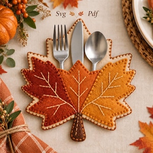 May include: A felt cutlery holder shaped like a maple leaf in shades of dark red, orange, and yellow, featuring cream-coloured blanket stitching along the edges and embroidered leaf veins. It holds a silver fork, knife, and spoon.