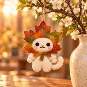Op de afbeelding: Een vilten kattenornament met een crèmekleurig lichaam en een grote, veelkleurige esdoornblad-hoofdtooi in tinten oranje, rood en groen. De kat heeft zwarte ogen, een klein geborduurd mondje en roze wangen, opgehangen aan een touwtje.