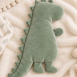 Puede incluir: Un cojín de peluche con forma de dinosaurio en color verde salvia y textura suave similar al terciopelo. El juguete relleno presenta una cabeza redondeada, una cola larga y una serie de púas triangulares en la espalda, con dos brazos y dos piernas.