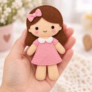 May include: A felt doll featuring brown hair with a pink bow, a pink dress with a white collar, and rosy cheeks. The doll has a simple stitched face with black eyes and a small smile, constructed from layered felt pieces with visible stitching.