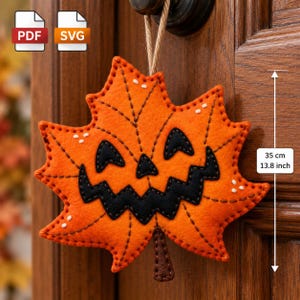 May include: An orange felt maple leaf decoration with a black stitched jack-o'-lantern face, hanging from a brown twine loop. The leaf features dark brown decorative stitching and measures 35 cm in total height as indicated.