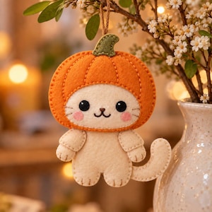 Op de afbeelding: Een hangend viltornament in de vorm van een crèmekleurige kat met een oranje pompoenhoed met een groene steel. De kat heeft zwarte geborduurde ogen, een klein neusje en roze wangen. Het heeft een bruin touwtje om het ornament op te kunnen hangen.
