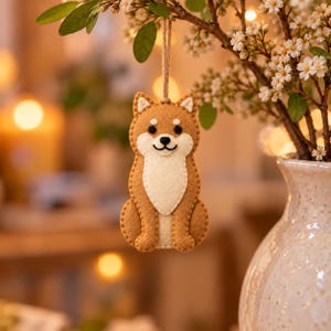 May include: A felt Shiba Inu dog ornament featuring a tan and cream color palette with stitched details, black eyes, and a black nose. The ornament is suspended by a thin brown twine loop, showcasing a sitting posture with a white chest patch.