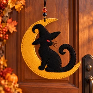 May include: A felt hanging ornament featuring a black cat wearing a pointed hat, sitting on a yellow crescent moon. The cat has red eyes and a curled tail. The ornament is suspended by a black string with red and black beads at the top.