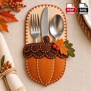 May include: An acorn-shaped felt cutlery holder in shades of orange and brown, featuring a decorative leaf and berry accent. The holder contains a stainless steel fork, knife, and spoon. The design includes visible stitching and PDF and SVG icons.