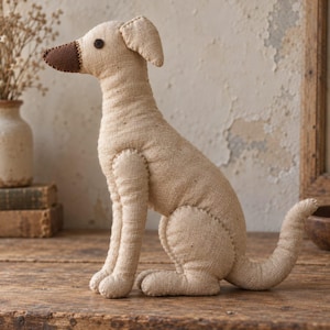 May include: A beige fabric dog figurine in a seated position, featuring a dark brown stitched nose and a small dark brown circular eye. The stuffed animal is constructed from a textured, natural-colored linen-like material with visible seams.