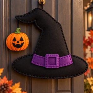 May include: A black felt witch hat wall hanging featuring a purple band with a square buckle and a small orange felt jack-o'-lantern pumpkin dangling from the brim by a black cord, all constructed with visible dark stitching along the edges.