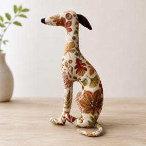 May include: A stuffed fabric dog figurine featuring a cream-coloured base with a printed pattern of autumn leaves in shades of orange, brown, yellow, and green. The dog has a dark brown ear, a button eye, and a small lace collar with a button detail.