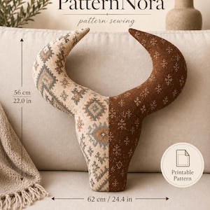 May include: A bull skull-shaped cushion featuring a split design with a cream-coloured side displaying a geometric pattern and a brown side with a snowflake-like print. The cushion measures 56 cm in height and 62 cm in width. Text reads PatternNora.