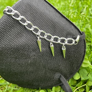 May include: A silver chain bracelet with three dangling, pointed cone charms. The bracelet is displayed on a black, textured surface against a backdrop of green grass and clover.