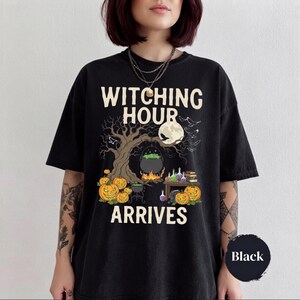 May include: A black t-shirt featuring the text "WITCHING HOUR ARRIVES" in cream lettering. The graphic depicts a large tree, a full moon with a bird, a bubbling cauldron over a fire, several jack-o'-lanterns, potion bottles, and books.