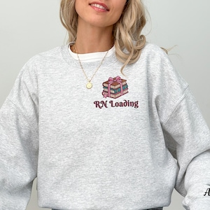 May include: Light gray sweatshirt with the text "RN Loading" and a graphic of stacked books tied with a pink bow. The sweatshirt is a crew neck style and has long sleeves. The color of the sweatshirt is ash.