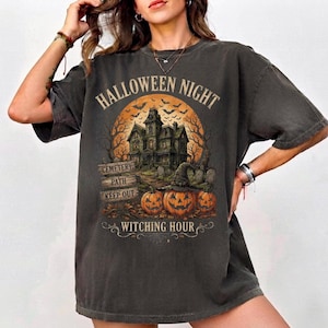 May include: A charcoal gray short-sleeve t-shirt featuring a graphic print of a haunted house under a large orange moon with bats. The design includes three jack-o'-lanterns, a sign reading CEMETERY PATH KEEP OUT, and the text HALLOWEEN NIGHT WITCHING HOUR.