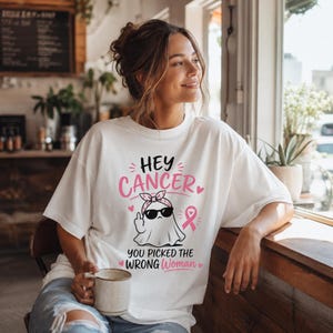 May include: A white t-shirt featuring a graphic of a ghost wearing a pink patterned headband and black sunglasses, giving a middle finger gesture. The text reads HEY CANCER YOU PICKED THE WRONG WOMAN, accompanied by a pink breast cancer awareness ribbon.