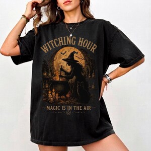 May include: A black short-sleeve t-shirt featuring a graphic print of a witch stirring a cauldron under a full moon. The text reads "WITCHING HOUR" at the top and "MAGIC IS IN THE AIR" at the bottom, accompanied by a black cat and candles.