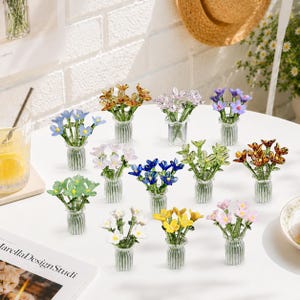 May include: A selection of miniature glass flower bouquets in various colours, including blue, yellow, pink, and orange. Each bouquet is displayed in a small, clear, ribbed glass vase. The flowers appear to be handcrafted.