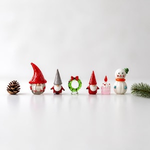 May include: A collection of miniature glass figurines including a brown pinecone, a gnome with a red hat, a gnome with a grey hat, a green wreath with a red bow, a gnome with a red hat, a small pink cake with a red candle, and a white snowman.