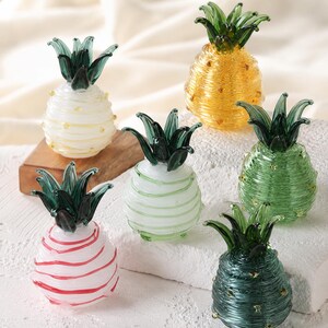 May include: A collection of six small glass pineapple figurines, each featuring a textured, spiraled body and a dark green glass leaf top. The pineapples are rendered in various colours, including white with yellow dots, solid yellow, light green, pink, and dark green.