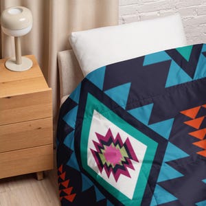 May include: A navy blue and teal geometric patterned blanket with orange and white accents. The blanket features a diamond-shaped design in the center with a pink and green floral pattern. The blanket is folded over a bed with a white pillow.