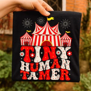 Puede incluir: Camiseta negra con un diseño de carpa de circo en rojo y blanco, y el texto "TINY HUMAN TAMER" en rojo y blanco. El diseño incluye fuegos artificiales y flores.
