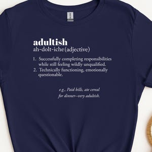 May include: Navy blue t-shirt with the word "adultish" and its definition printed in white. The definition includes two points and an example sentence. The shirt is knotted at the bottom.