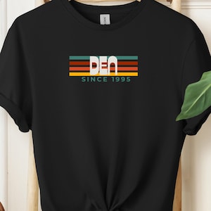 May include: A black short-sleeve t-shirt featuring a graphic on the chest with horizontal stripes in teal, orange, and yellow. The text "BEN" is printed in large white letters over the stripes, with the words "SINCE 1995" in smaller white text below.