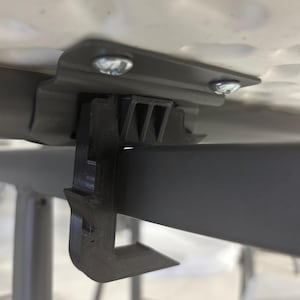 May include: Close-up of a black 3D-printed table clip attached to a gray metal table frame. The clip is secured to the table with two silver screws. The table surface is white with a textured pattern.