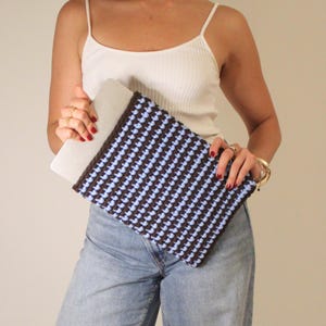 May include: A brown and light blue crocheted laptop sleeve. The sleeve features a repeating pattern of light blue squares against a brown background. The laptop sleeve is held by a person wearing a white vest top and blue jeans.
