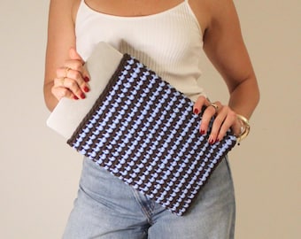 May include: A brown and light blue crocheted laptop sleeve. The sleeve has a repeating pattern of light blue squares against a brown background. The laptop sleeve is held by a person wearing a white tank top and blue jeans.