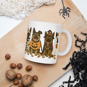 以下が含まれることがあります： ミイラ、カボチャ、魔法使い、幽霊のハロウィーンの衣装を着た4匹のジャーマンシェパード犬のカラフルなイラストが描かれた白いセラミックマグカップ。イラストのベース部分には秋の落ち葉が散りばめられています。
