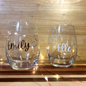Op de afbeelding: Twee heldere, stelenloze wijnglazen. Eén glas heeft de naam "Emily" in een sierlijk lettertype met een bloemenontwerp. Het andere glas heeft de naam "Elle" in een sierlijk lettertype met een vlinderontwerp. Beide glazen worden verlicht door feeërieke lichtjes.