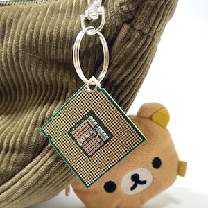 Könnte beinhalten: Ein einzigartiger Schlüsselanhänger mit einem Computerprozessor-Chip mit einem goldenen Raster und einem braunen Zentrum, befestigt an einem silbernen Schlüsselring und Verschluss. Ein kleiner, plüschiger Bärenanhänger ist ebenfalls befestigt.