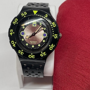 以下が含まれることがあります： 丸い文字盤と黒い穴あきバンドが付いた黒いSwatchの腕時計。文字盤の中央は薄茶色で、黒と黄色のマーキングがあります。白いスタンドに、赤い布の隣に飾られています。