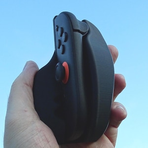 Puede incluir: Un mando de juegos portátil negro con un botón rojo y varios botones negros. El mando se sostiene en una mano sobre un fondo de cielo azul. El mando parece estar plegado.