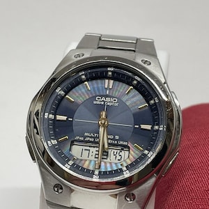 Pode incluir: Um relógio Casio Wave Ceptor prateado com mostrador azul e visor digital mostrando a hora. O relógio tem uma pulseira de aço inoxidável e um mostrador redondo com marcadores de minutos. O mostrador do relógio também tem o texto "CASIO wave ceptor".