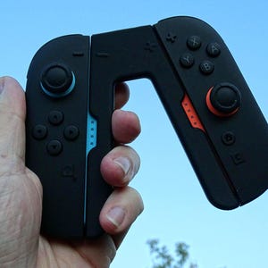 Può includere: Controller di gioco portatile nero con dettagli blu e arancioni. Il controller ha pulsanti, pad direzionali e levette analogiche. Il dispositivo è tenuto contro un cielo blu.