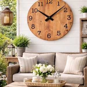 May include: A large, round wooden clock, 42 inches in diameter, with black numerals and hands, hangs on a white wall. The clock is part of an outdoor living space with a sofa, pillows, and a coffee table. The text on the wall says "Assembly required."
