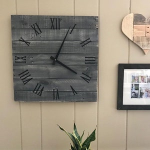 K&ouml;nnte beinhalten: Eine graue Holzuhr mit r&ouml;mischen Ziffern an einer wei&szlig;en Wand. Die Uhr hat schwarze Zeiger und ist von einem Bilderrahmen und einem Holzhertz umgeben.