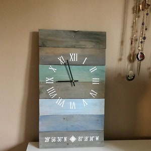 May include: A wooden clock with Roman numeral markings and a compass design. The clock face is painted in shades of blue and green, with a white background. The clock is mounted on a wall and has a white background.