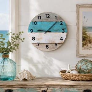 May include: A round, wooden wall clock with a coastal theme. The clock face features horizontal stripes in gray, teal, and white. Black hands indicate the time. The clock is mounted on a white wall, with beach-themed decor in the foreground.