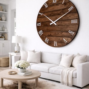 May include: Large, round wooden wall clock with white Roman numerals and hands. The clock is mounted on a white wall above a white sofa. The clock's dark brown wood grain contrasts with the white interior decor.