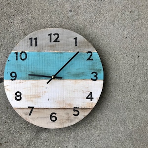 May include: A round wooden clock with black numbers and hands. The clock face is painted in stripes of white, blue, and white. The clock is hanging on a gray wall.