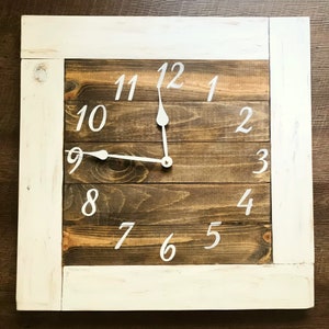 May include: A white framed wooden clock with white painted Roman numerals on a brown wood background.