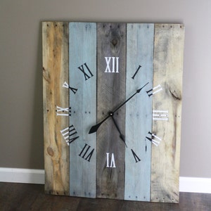 May include: A large, square wooden clock with a rustic design. The clock face is made of vertical wooden planks in shades of blue, gray, and natural wood tones. Black clock hands and white Roman numerals mark the time. The clock measures approximately 24 inches by 24 inches.