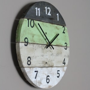 May include: A round, rustic wall clock made of distressed wood. The clock face is divided into three horizontal sections: dark gray, light green, and off-white. Black numerals mark the hours, and black hands indicate the time. The clock has a farmhouse style.