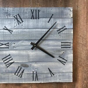 K&ouml;nnte beinhalten: Eine graue Holzuhr mit r&ouml;mischen Ziffern und schwarzen Zeigern. Die Uhr ist aus wiederverwendeten Holzbrettern gefertigt.
