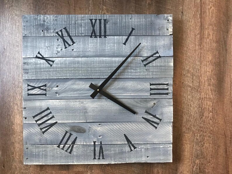 K&ouml;nnte beinhalten: Eine graue Holzuhr mit r&ouml;mischen Ziffern und schwarzen Zeigern. Die Uhr ist aus wiederverwendeten Holzbrettern gefertigt.